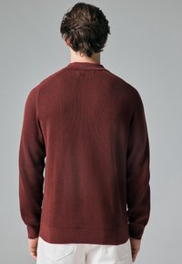 Pull en tricot de couleur rouge profond, avec une texture côtelée, des manches longues et un col rond, vu de dos sur un fond uni.