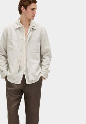 Mann trägt eine beige Jacke mit Knöpfen über einem hellen Hemd und braune Hosen, Hände in den Taschen, schaut nach links vor einem einfarbigen Hintergrund.