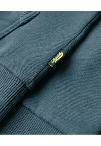 Acercamiento a una tela de punto en color azul verdoso, con puño acanalado y una pequeña etiqueta verde con un logo amarillo.