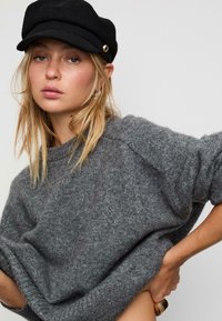 Maglione grigio a maglia con una vestibilità morbida e polsini a coste, abbinato a un berretto di lana nero con un accento di bottone dorato. Texture morbida evidente.