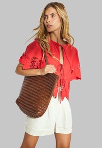 Blusa rossa ricamata a maniche corte, abbinata a pantaloni corti di lino beige. Tiene una borsa a tracolla in tessuto intrecciato di colore marrone con motivo a spina di pesce.