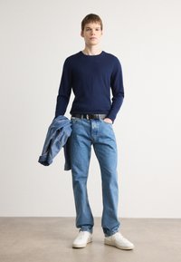 Pulover tricotat bleumarin, blugi deschiși din denim, adidași albi și o jachetă din denim purtată pe braț, stând în fața unui fundal neutru.