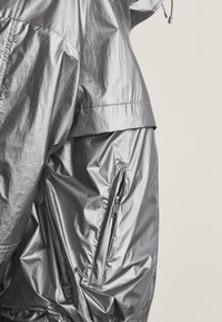 Nahaufnahme einer Person, die eine glänzende silberne Jacke mit einer Reißverschlusstasche an der Seite und einer sichtbaren Kordelzugkapuze trägt.