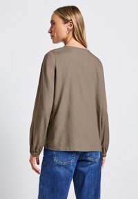 Blusa marrone a maniche lunghe con scollatura rotonda e polsini arricciati; abbinata a pantaloni in denim blu con tasche posteriori.