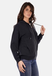 Felpa nera con zip e cappuccio, caratterizzata da una superficie liscia, tasche laterali e una cintura aderente. Il modello tiene il cordino.