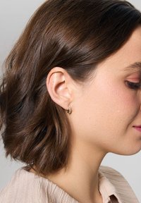 Złota kolczyk w kształcie obręczy z teksturowaną powierzchnią, ozdobiony małymi błyszczącymi akcentami. Noszony na lewym uchu, prezentujący widok z bliska.