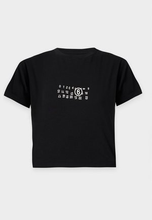 Kobieta w czarnym krótki t-shircie, długiej dwukolorowej jeansowej spódnicy, czarnych botkach, trzymająca małą czarną torebkę, stoi na białym tle.
