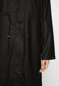 Abrigo negro impermeable con un diseño elegante, cordones ajustables, botones de presión y una textura suave y brillante.