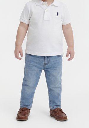 Niño pequeño vistiendo una camisa polo blanca de manga corta, vaqueros azul claro y zapatos náuticos de cuero marrón, de pie contra un fondo blanco.