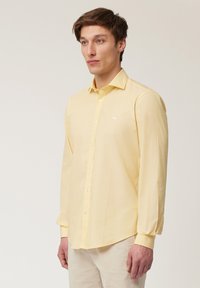 Camisa amarilla de manga larga con cuello abotonado y un pequeño logo blanco en el pecho. Hecha de una tela ligera y suave.