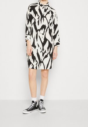 Persoon draagt een zwart-wit abstracte print jurk in overhemdstijl, gecombineerd met witte sokken en zwarte hoge sneakers, staande tegen een effen achtergrond.