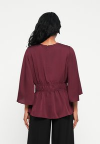 Blusa bordeaux in tessuto leggero e fluido. Presenta una vita arricciata, maniche a campana e chiusura con zip sul retro. Nessun ulteriore dettaglio.