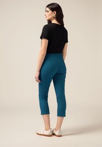 Pantaloni cropped teal con tasche posteriori, abbinati a una maglietta nera a maniche corte e sneakers crema. Tessuto liscio, design aderente.
