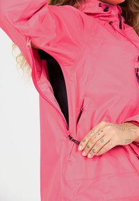 Personne portant une veste rose vif déboutonnant l’ouverture de ventilation sous le bras, montrant la doublure en mesh et une main tatouée ajustant la fermeture éclair.