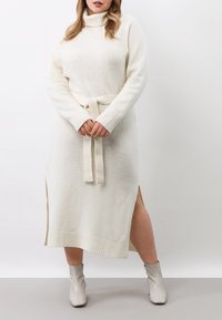 Lång, krämfärgad stickad klänning med hög krage, bälte i midjan, sidslitsar och ribbade ärmslut. Bärs med ankelboots.