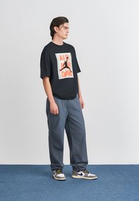 Μαύρο oversized t-shirt με λευκή και πορτοκαλί εκτύπωση γραφικών, συνδυασμένο με χαλαρούς γκρι παντελόνες και λευκά παπούτσια με μαύρες λεπτομέρειες.