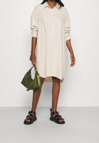 Helles beige Leinenhemdkleid mit Knopfleiste und langen Ärmeln. Schwarze Riemchensandalen und eine kleine grüne Satintasche, die in einer Hand gehalten wird.