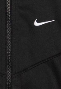 Jaqueta preta com fecho de correr, apresentando um logótipo da Nike em branco contrastante. O tecido possui uma textura suave com detalhes de costura angulada.