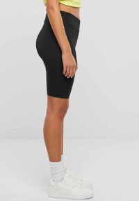 Schwarze, dehnbare Radlerhosen mit hohem Bund, glatter Textur und eng anliegendem Design. Kombiniert mit weißen Sneakers und Knöchelsocken.