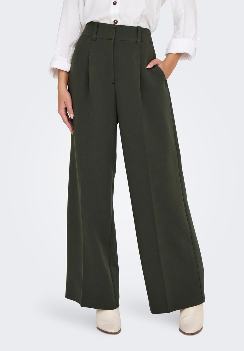 Donna che indossa pantaloni verde scuro a gamba larga, camicia bianca con bottoni e maniche arrotolate, e stivaletti bianchi. Mani in tasca.