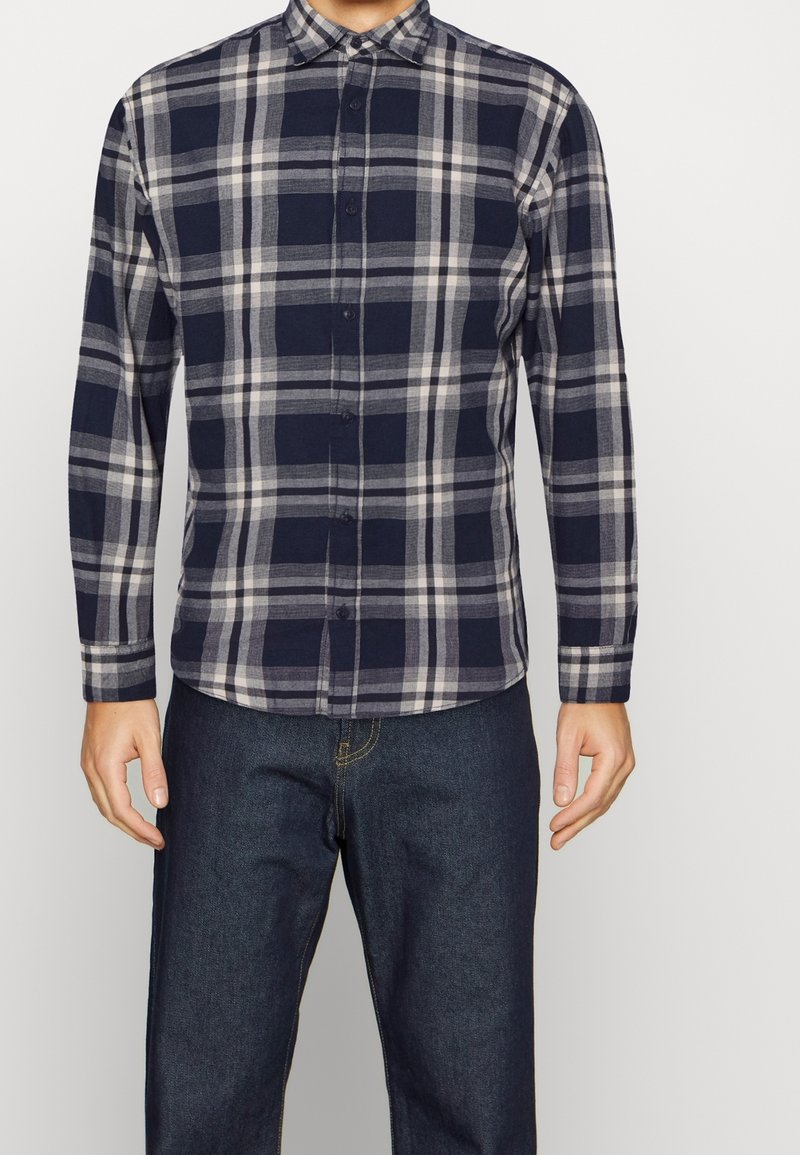 Mörkblå och grå rutig skjorta med lång ärm, knappkrage och knappar framtill. Tyget verkar mjukt och texturerat. Bärs med blå jeans.