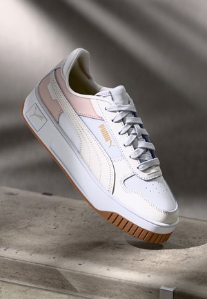 Baskets Puma blanche et rose clair avec semelle en gomme et lacets, flottant au-dessus d'une surface en béton gris avec un arrière-plan d'ombre douce.