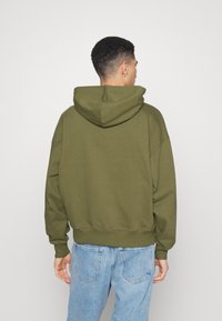 Kaki hettegenser med stor hette og avslappet passform. Har ribbestrikkede mansjetter og nederkant, kombinert med lyseblå jeans. Enkel design, ingen synlige logoer.
