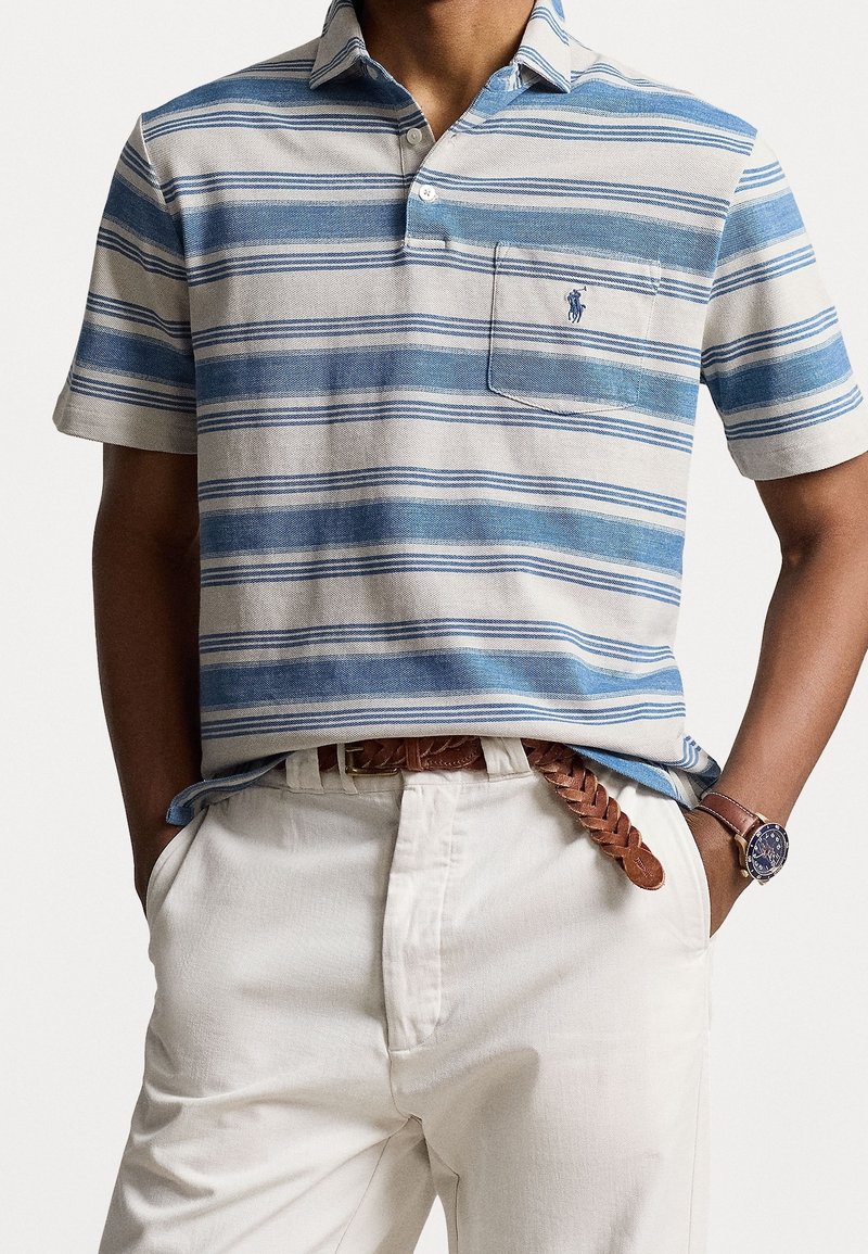 Homme portant un polo rayé bleu et blanc avec une poche poitrine et un pantalon blanc avec une ceinture tressée marron et une montre-bracelet.