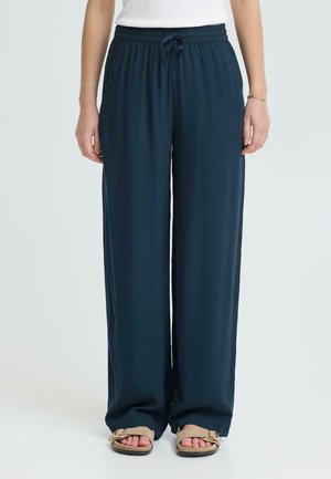 Kobieta w granatowych szerokich spodniach z troczkiem i beżowych sandałach z odkrytymi palcami, stojąca na tle jasnego, jednolitego tła.