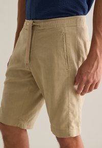 Shorts en coton beige à coupe droite, avec un cordon de serrage, des poches latérales et des ourlets repliés pour une finition soignée.