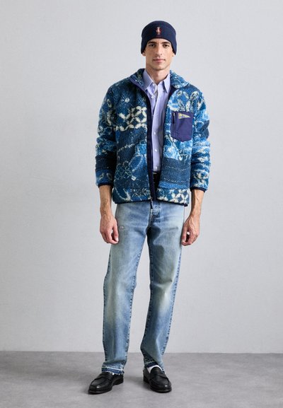 Flauschige Jacke in Blau- und Weißmuster mit einer dunkelblauen Brusttasche, hellblauem Hemd, ausgewaschenen blauen Jeans und schwarzen Slippern.