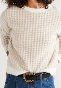 Helle Cremefarbene Strickjacke mit einem strukturierten Hahnentrittmuster, ausgestattet mit einem runden Ausschnitt, kombiniert mit blauer Jeans und einem schwarzen Gürtel mit einer goldenen Kettenschnalle.