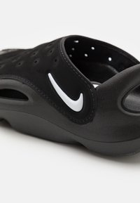 Black slide sandal made of textured synthetic material. Features ventilation holes, a white Nike logo, and a contoured design for comfort.
