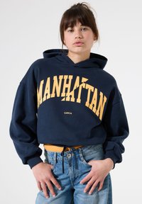 Jeune femme avec les cheveux bruns en chignon, portant un hoodie bleu marine avec le texte "MANHATTAN" et un jean bleu clair, se tenant devant un fond clair uniforme.