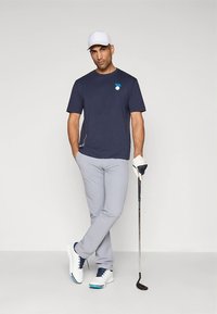 Mørkeblå golfskjorte med korte ermer og liten logo, lysegrå smale bukser, hvite golf-sko med blå detaljer, holder en golfkølle.