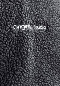 Tissu en polaire noir texturé avec texte brodé blanc "Originals Studio" centré sur le matériau.