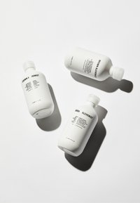 Trois bouteilles en plastique blanc de produits capillaires avec des étiquettes en texte noir. Les bouteilles sont cylindriques avec des bouchons à rabat, affichant divers noms de produits.