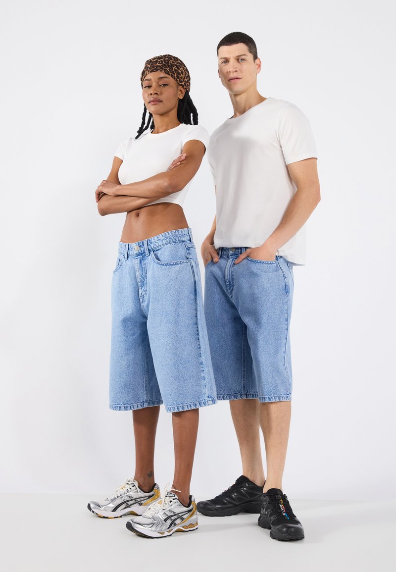 Two models wearing casual outfits: a fitted white T-shirt and light blue denim shorts. The shorts have a relaxed fit and frayed hems.