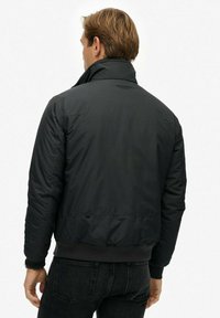Chaqueta acolchada negra con un cuello alto, puños acanalados y una cintura ajustada. Tejido texturizado, diseñado para usar en invierno. Sencilla y funcional.