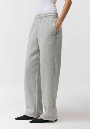 Osoba ubrana w luźne szare spodnie dresowe z elastycznym pasem i czarne szpiczaste buty, stojąca z jedną ręką w kieszeni.