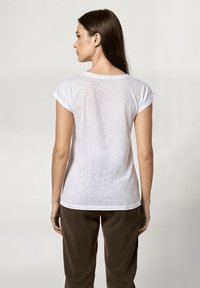 Camiseta blanca de manga corta con un escote redondeado, fabricada en un tejido ligeramente transparente. Corte holgado que llega a las caderas, combinada con pantalones marrones.