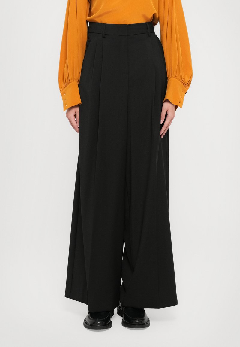 Pantaloni neri a gamba larga con vita alta, dettaglio plissettato nella parte anteriore e texture liscia, abbinati a una blusa arancione a maniche lunghe.