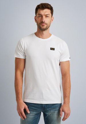 Camiseta de algodón blanca de manga corta, con cuello redondo y un pequeño parche con logotipo negro y dorado en el lado izquierdo del pecho. Diseño simple y sólido.