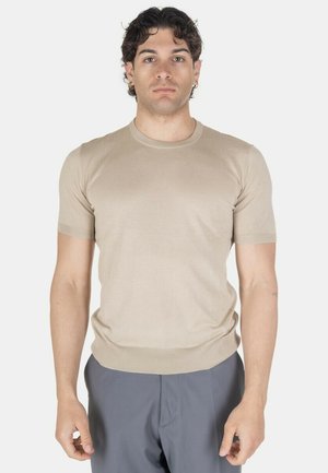 Giovane uomo con capelli corti, ricci e scuri indossa una maglietta beige a maniche corte in maglia e pantaloni grigi, in piedi di fronte a uno sfondo bianco.