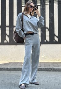 Una donna in tuta grigia coordinata e occhiali da sole si posa per un selfie in strada, portando una grande borsa a tracolla marrone.