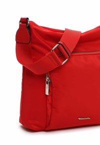 Punane nailonist õlakott laiendatava õlarihmaga. Esiküljel on tõmblukuga tasku ja metallist brändi logo detail.
