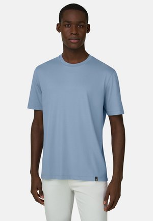 Man draagt een lichtblauw T-shirt met korte mouwen en witte broek, staand tegen een effen witte achtergrond.