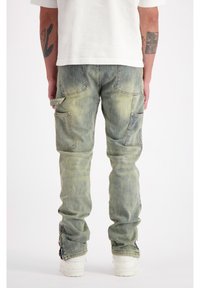 Vervaagde blauw-groene denim cargobroek met meerdere zijzakken, lichte slijtage en taps toelopende benen, gedragen met witte sneakers.
