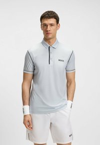 Polo shirt grigio chiaro con maniche corte, decorato con strisce nere e ambra, con un logo sul petto. Tessuto morbido, design classico.