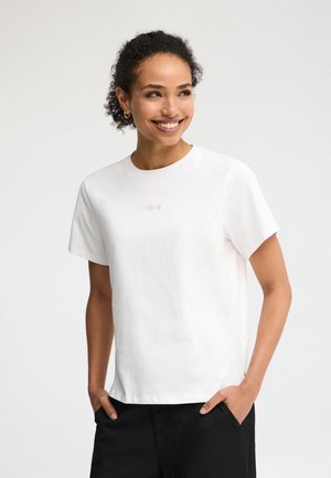 Lächelnde Frau mit lockigem Haar, die goldene Creolen, ein weißes Kurzarmshirt und schwarze Hosen trägt, Hände in den Taschen vor neutralem Hintergrund.